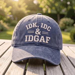 Puede incluir: Una gorra de béisbol azul con acabado desgastado que presenta texto bordado en blanco que dice IDK, IDC & IDGAF. La gorra tiene una visera curva, un botón en la parte superior y ojales cosidos en la corona, con un diseño estructurado.