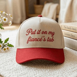 Könnte beinhalten: Eine Baseballkappe mit cremefarbener Krone und rotem Schirm. Auf der Vorderseite der Krone befindet sich der in roter Schreibschrift gestickte Text "Put it on my fiancé's tab". Die Kappe hat ein strukturiertes Design mit fünf Paneelen.