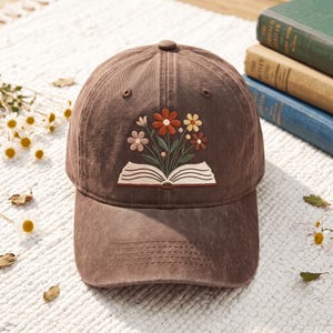 Op de afbeelding: Een bruine baseballpet met een borduursel van een open boek waaruit een boeket bloemen in rood-, geel- en rozetinten groeit. De pet heeft een gestructureerde kroon, een gebogen klep en een knoopje aan de bovenkant.