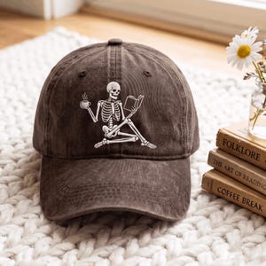 Puede incluir: Una gorra de béisbol de color marrón oscuro con un gráfico blanco de un esqueleto sentado que sostiene una taza de café humeante en una mano y un libro abierto en la otra. La gorra tiene una textura desgastada y una visera curva.
