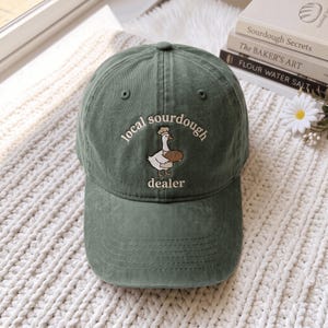 Può includere: Un cappellino da baseball verde salvia con il ricamo di un'oca bianca che indossa un cappello da chef e tiene in mano una pagnotta marrone. Il testo "local sourdough dealer" è ricamato in bianco ad arco sopra e sotto l'oca.