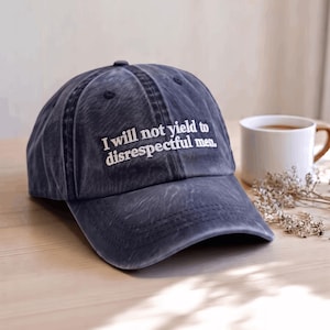 May include: A charcoal grey baseball cap with a distressed, washed-out finish. The front panel features white embroidered text that reads, "I will not yield to disrespectful men." The cap has a curved brim and a structured crown with ventilation eyelets.