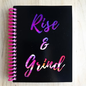Op de afbeelding: Een zwarte spiraalgebonden notitieboek met de woorden "Rise & Grind" in een kleurverloopschrift. De woorden zijn gedrukt op een zwarte achtergrond met een kleurverloopontwerp.
