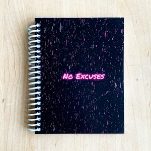 May include: A black spiral bound notebook with a pink and black speckled design. The notebook has the words "NO EXCUSES" in pink neon lettering.