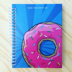 May include: A blue and pink donut notebook with a white spiral binding. The donut has pink icing with colourful sprinkles and a blue centre. The notebook has the text "www.trainrite.ca" printed on the front cover.