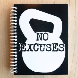 May include: A spiral-bound notebook with a black cover featuring a white kettlebell graphic and the words "NO EXCUSES" in bold, distressed font. The notebook is open, revealing blank pages, and rests on a light wood surface.