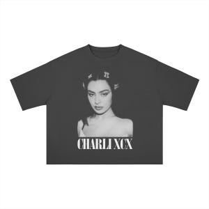 Pode incluir: Uma t-shirt de manga curta cinzento-escura com uma impressão fotográfica a preto e branco de uma pessoa com ganchos no cabelo na zona do peito. Abaixo da imagem, o texto CHARLI XCX está impresso numa fonte serifada branca e negrito.