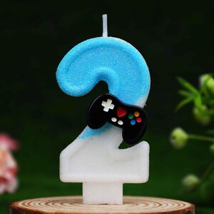 May include: A number two birthday candle featuring a blue glittery top half and a white glittery bottom half. A small black video game controller icon with a white directional pad, a red heart button, and four colored action buttons is attached.
