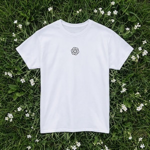 Op de afbeelding: Een wit T-shirt met korte mouwen en een klein, zwart, cirkelvormig geometrisch logo in het midden op de borst. Het shirt heeft een klassieke ronde hals en een rechte zoom, gemaakt van effen witte stof met een strak ontwerp.