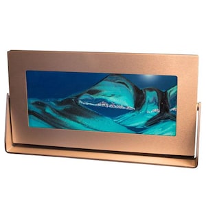 May include: A rectangular sand art display featuring a metallic rose gold frame and stand. The central glass panel contains layers of vibrant turquoise, deep blue, and black sand, which are arranged to form abstract mountain-like peaks and valleys.