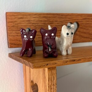 May include: Three small cat figurines standing on a wooden shelf. Two of the cats are dark maroon with white dot eyes and stitched details, while the third cat is a shimmering off-white color with dark eyes and a small horn on its head.