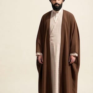 May include: A person wearing a dark brown open-front robe over a light beige long-sleeved tunic. The person has a dark beard and wears a black turban on their head. The outfit features clean lines and a layered appearance with a neutral color palette.
