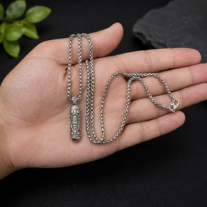 Puede incluir: Un colgante cilíndrico de tono plateado con símbolos grabados, suspendido de una cadena de eslabones cuadrados a juego. El colgante tiene extremos redondeados con patrones de puntos decorativos y la cadena incluye un cierre de mosquetón.