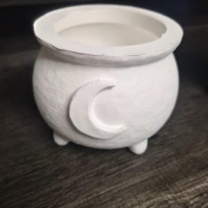 May include: A small, round, white cauldron-shaped pot featuring a raised crescent moon emblem on the front. The vessel has a wide, open top rim and is supported by three short, rounded feet, with a textured surface finish throughout the exterior.
