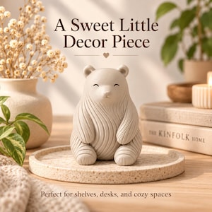 May include: A beige bear figurine with a smooth, matte finish and textured, swirling carved lines across its body. The bear is seated in a forward-facing position on a round, speckled, light-colored tray. The text A Sweet Little Decor Piece is visible.