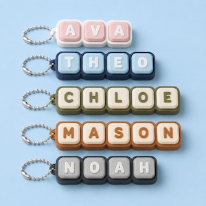 May include: Five rectangular keychains with metal ball chains, each featuring raised block letters spelling the names AVA, THEO, CHLOE, MASON, and NOAH. The keychains have coloured frames and white or coloured square tiles with embossed letters.