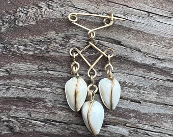 May include: A gold-toned metal brooch with a safety pin closure and three white shell-like pendants hanging from it.