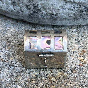 May include: A small, rectangular silver box with iridescent abalone shell inlays. The box has a hinged lid and a clasp closure. The box is sitting on a textured, gray surface.