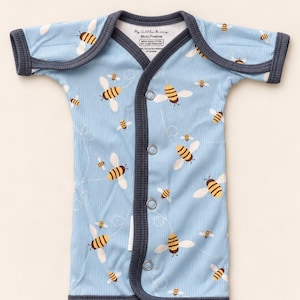 Puede incluir: Un body de manga corta azul claro con textura acanalada y un estampado de abejas amarillas y negras con alas blancas. La prenda tiene ribetes gris oscuro, cierre frontal con botones a presión y una etiqueta que dice My Little Bunny Micro Preemie.