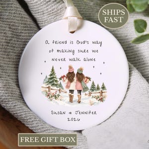 May include: A round white ceramic ornament with a cream ribbon hanger. The design features two people walking in a snowy landscape with pine trees and a fence. Text reads: A friend is God's way of making sure we never walk alone, Susan & Jennifer, 2026.