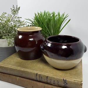 May include: Two small ceramic crocks with a two-tone glaze. The left crock is dark brown with a tan rim, while the right crock features a dark brown upper section and a tan lower section with small side handles. Both are resting on old books.