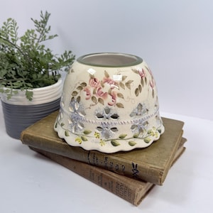 May include: A cream-colored ceramic candle holder with a scalloped base and cut-out floral patterns. The exterior features hand-painted pink roses, green leaves, and small yellow flowers. A thin green band circles the top rim of the object.