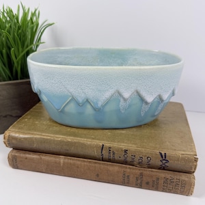 May include: A ceramic bowl with a light blue base and a textured white glaze dripping down from the rim in a mountain-like pattern. The bowl is resting on two stacked vintage books with visible text on the spines, including the title Essentials of Arithmetic.