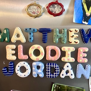 May include: A collection of ceramic alphabet magnets arranged to spell the names Matthew, Elodie, and Jordan. Each letter features unique patterns including stripes, polka dots, stars, flowers, clouds, and trees in various colors.