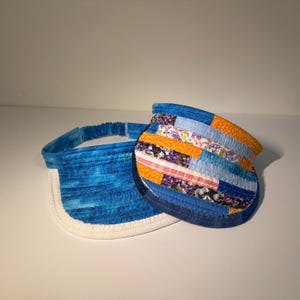 Può includere: Due visiere in tessuto con cuciture trapuntate. Una visiera presenta un motivo blu marmorizzato con la parte inferiore bianca. La seconda visiera mostra un design patchwork di strisce di tessuto arancione, azzurro e floreale con bordo blu scuro.