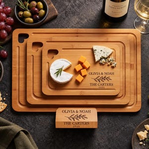 May include: A set of three rectangular bamboo serving boards with heart-shaped cutouts and a matching stand. The boards and stand are engraved with the text OLIVIA & NOAH, THE CARTERS, and 2025, accompanied by a leafy branch illustration.