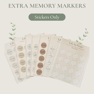 May include: A set of sticker sheets titled EXTRA MEMORY MARKERS and Life In Firsts. The sheets feature circular labels with text such as First Smile, First Steps, First Kiss, First Concert, and High School Graduation, arranged in columns on a light background.