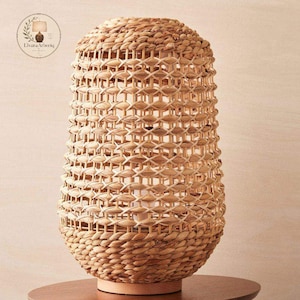 Pode incluir: Um candeeiro de mesa com um abajur de fibras naturais entrançadas com um padrão de rede aberto. A base é feita de madeira de cor clara e a estrutura é cilíndrica com um topo ligeiramente afunilado em tons de bege e castanho claro.