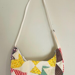 May include: A crescent-shaped hobo bag constructed from various patchwork fabric pieces. The exterior features a mix of floral, polka dot, and striped patterns in yellow, red, green, and grey. It includes a long, solid white fabric shoulder strap.