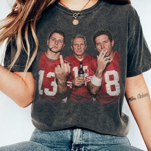 May include: A charcoal grey short-sleeve t-shirt featuring a graphic print of three individuals in red football jerseys with the numbers 13, 16, and 8. The graphic shows the individuals smoking and holding a beer bottle, with one person gesturing.