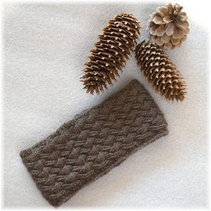 May include: A brown knitted headband with a cable knit pattern. The headband is lying on a white surface with pine cones.