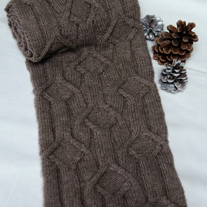May include: A brown knitted scarf with a cable knit pattern. The scarf is folded in half and lying on a white surface.