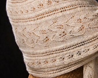 May include: A cream-coloured knitted shawl with detailed openwork and lace patterns. The shawl is draped over the shoulders, highlighting the delicate texture. The design features horizontal stripes and floral motifs, creating an elegant and airy appearance. The shawl is made of a soft, light yarn.