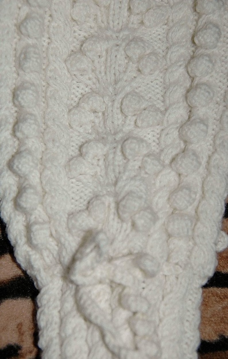 Może przedstawiać: Zbliżenie kremowej dzianiny o wzorze strukturalnym. Wz&oacute;r charakteryzuje się wypukłymi, zaokrąglonymi bobbles i pionowymi sekcjami warkoczowymi. Tkanina wydaje się miękka i ciepła, odpowiednia na sweter lub koc.