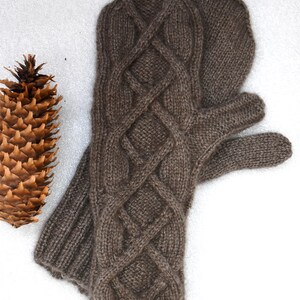 May include: A pair of brown knitted mittens with a cable knit design. The mittens are made of a soft, textured yarn and have a ribbed cuff. A pine cone is visible in the background.