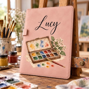 Könnte beinhalten: Ein rechteckiger, hellrosa Notizbuchumband mit dem Namen Lucy in schwarzer Schreibschrift. Unter dem Namen befindet sich eine Aquarellillustration eines offenen Malkastens mit zwölf Farbnäpfchen, einem Pinsel und weißen Blütenzweigen.