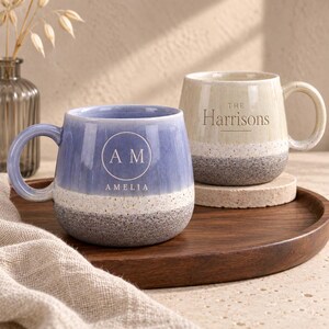 Peut inclure: Deux tasses en céramique avec anses. La tasse avant est bleue avec une bande blanche tachetée et une base grise tachetée, portant un logo circulaire avec les lettres AM et le nom AMELIA. La tasse arrière est crème avec une base grise tachetée et le texte THE Harrisons.