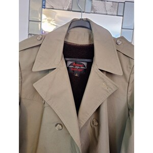 May include: A tan double-breasted trench coat with epaulets on the shoulders and a dark brown interior lining. The inner label reads Botany Weather Topper 42 LONG. The coat features wide lapels, front buttons, and a structured collar design.
