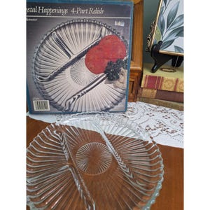 May include: A clear glass 4-part relish tray with a radial ribbed pattern and a central circular design. The tray is displayed in front of its original blue and white cardboard box labeled Crystal Happenings 4-Part Relish.