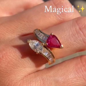 May include: A bypass ring featuring a pear-shaped red gemstone and a pear-shaped clear gemstone, each accented by a row of baguette-cut clear stones set in a gold-toned band. The word Magical is printed in white text with two yellow star icons.