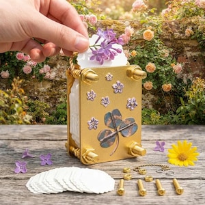 May include: A gold-colored metal flower press featuring a decorative clover emblem and small floral accents on the front plate. The device includes threaded metal rods, nuts, a stack of white scalloped paper inserts, and a small metal chain.