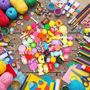 May include: A collection of colourful craft supplies including yarn balls, beads, paint tubes, coloured pencils, scissors, and various small candy-shaped resin charms arranged on a wooden surface. The charms feature shapes like lollipops, donuts, and sweets.