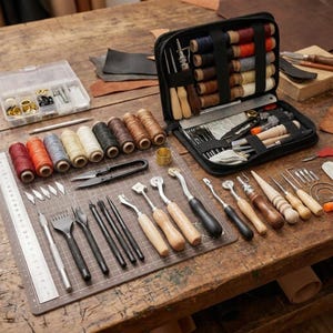 Könnte beinhalten: Ein umfassendes Lederbearbeitungsset mit einem offenen schwarzen Reißverschlussetui voller Garnrollen und Metallwerkzeuge sowie einer Schneidematte mit einer Auswahl an Stechbeiteln, Ahlen, Rillern und Metalllinealen.