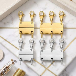 May include: Four rectangular magnetic jewelry clasps in gold and silver tones. Two clasps feature two lobster claw attachments, while two feature three lobster claw attachments. Each clasp consists of two interlocking metal bars with loops.