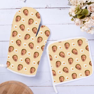 May include: A kitchen oven mitt and a square pot holder set in a light beige color. Both items feature a repeating pattern of a man's face with an open mouth and a small fried egg graphic. The fabric accessories are designed for heat protection.