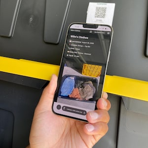 May include: A smartphone displaying a Notion page titled Wife's Clothes with a QR code tag labeled GT-001 visible above the device. The screen shows a digital inventory list including location details, a photo of folded clothing, and a tag ID.